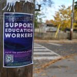 Photograph of a streetscape. There is a wooden hydro pole in focus on the left of the photo. Pole has a purple poster on it that says "Support Education Workers" with smaller white text below that says "Paint the province purple!" and smaller text underneath. The autumn streetscape in the background is out of focus, showing a road, a crosswalk, and trees with yellow leaves