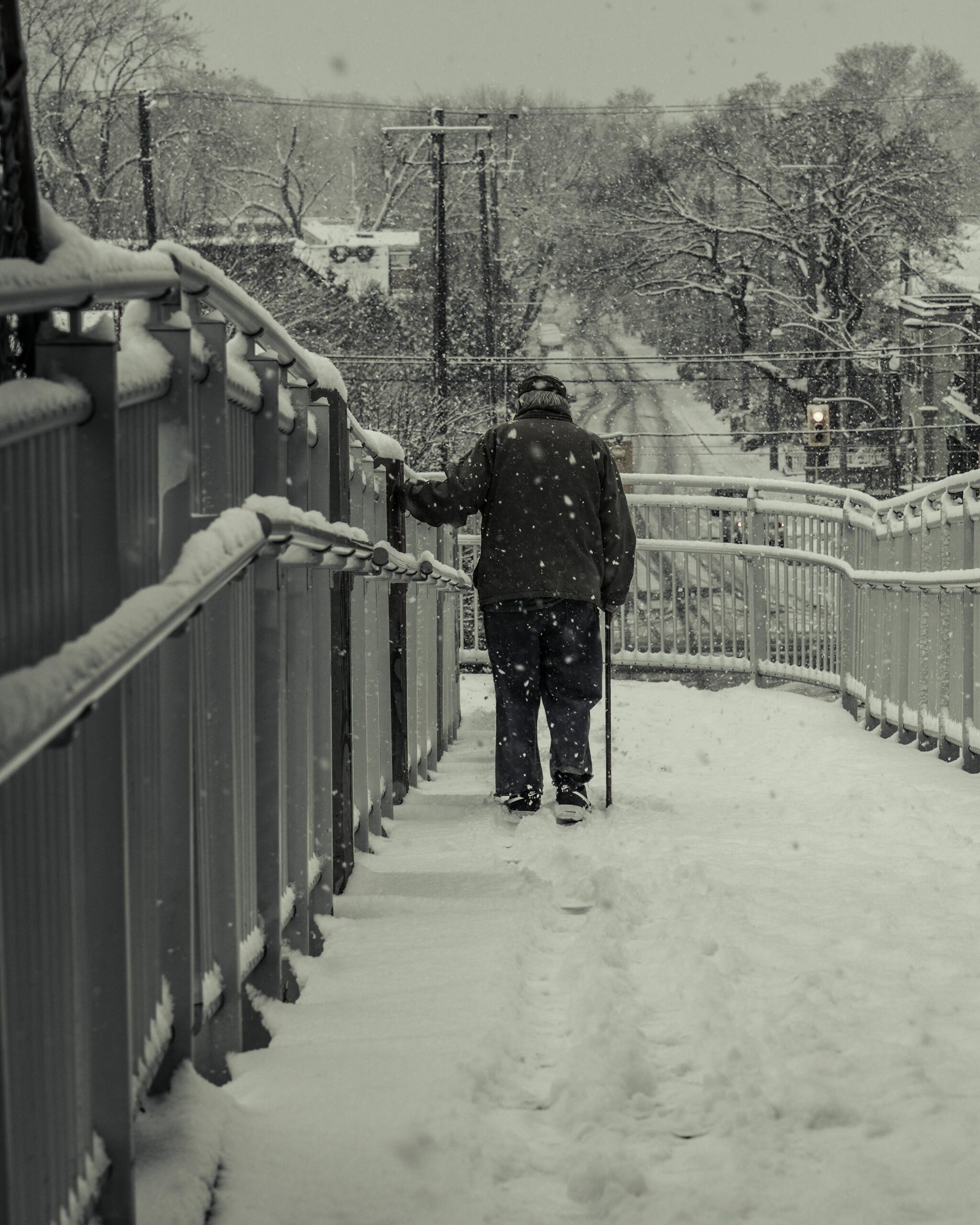 Grey, white and black photo of older gentleman walking away from the viewer, over a bridge in the snow, with his left hand on the railing, using a cane with his right hand. Photo by The Ahnaf Piash via pexels